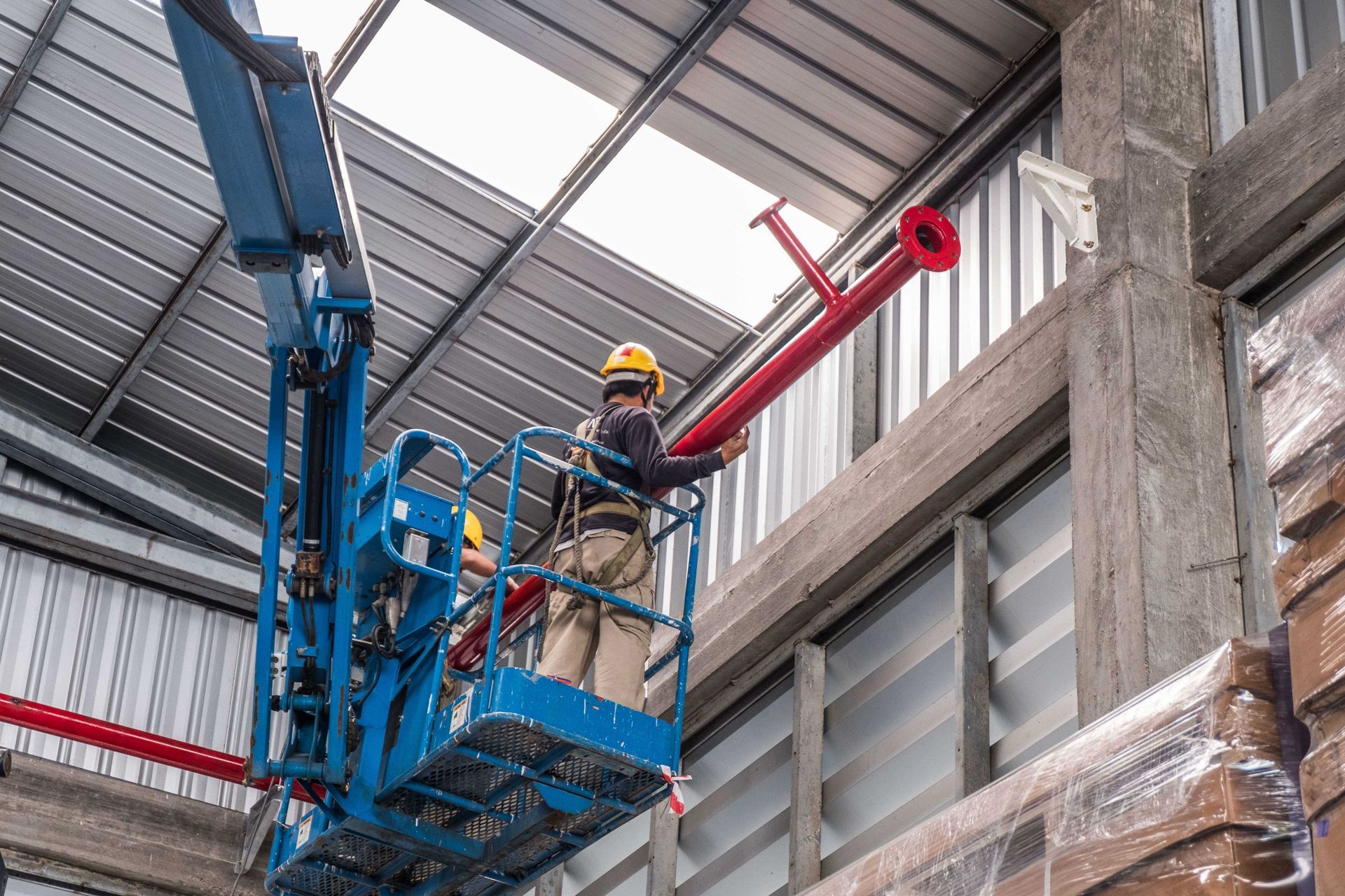 Worker in yellow hard hat on a blue lift inside a warehouse, near stacked materials and metal walls