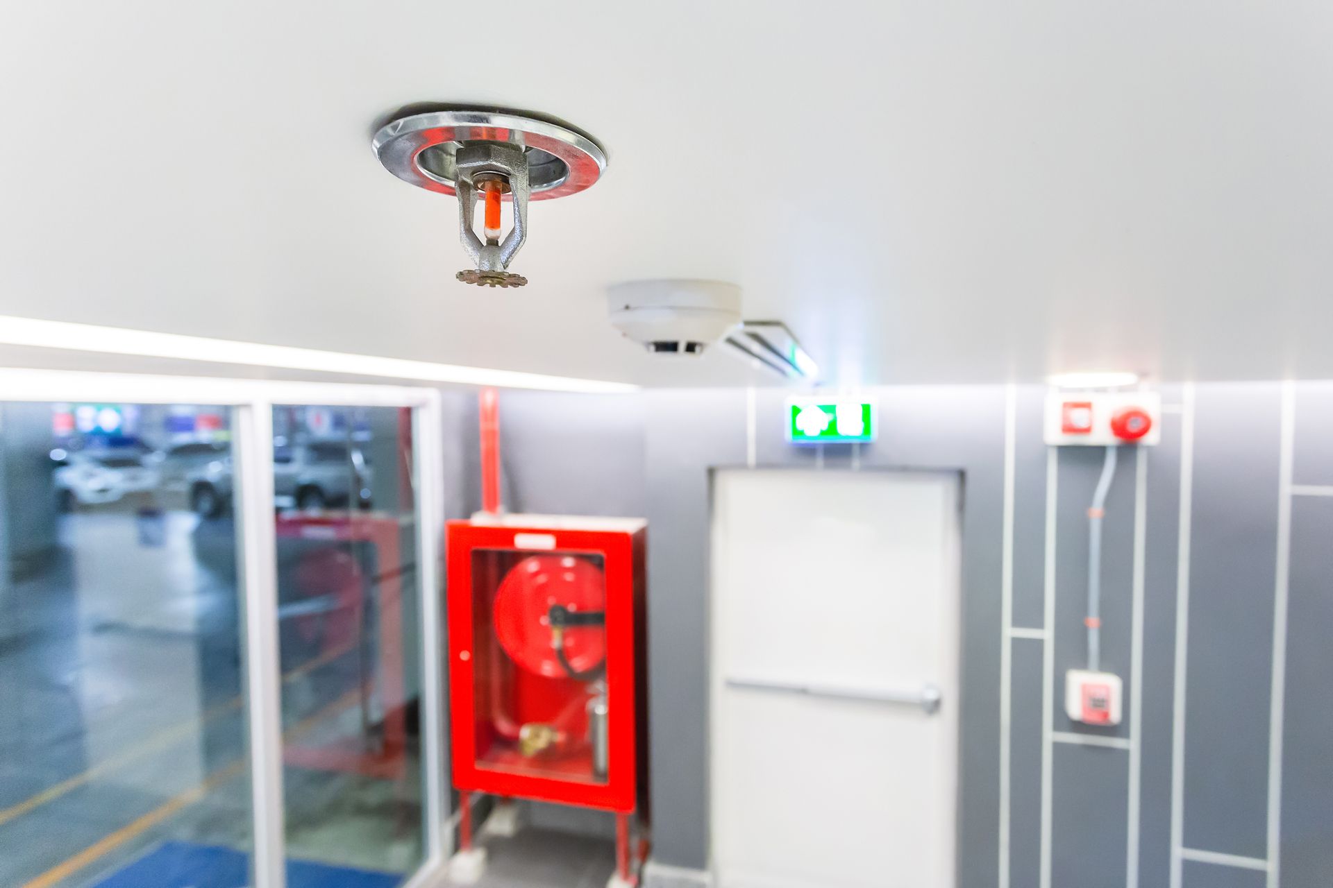 Ceiling-mounted fire sprinkler and smoke detector above a white door, red fire cabinet, and exit signs in a hallway