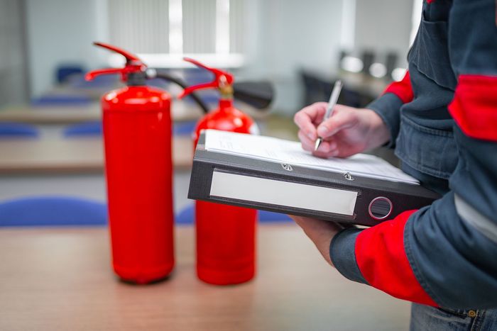 Person writing on a clipboard beside red fire extinguishers in an indoor setting