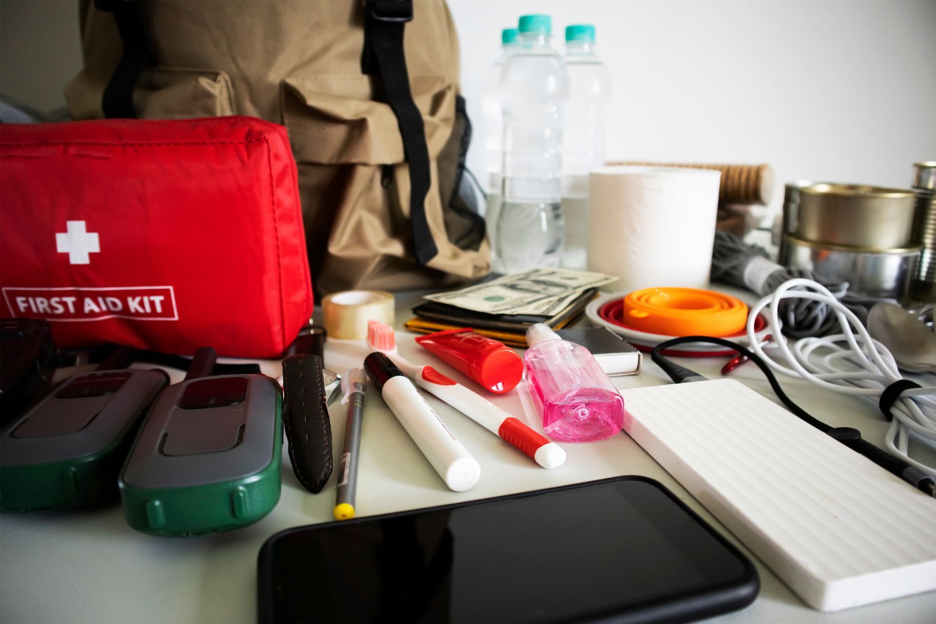 Emergency supplies and gadgets spread on a table, with a red first aid kit and water bottles.