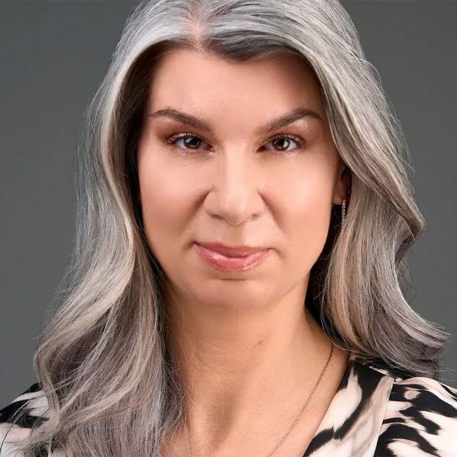 Woman with long gray hair, wearing a patterned top, looking directly at the viewer.