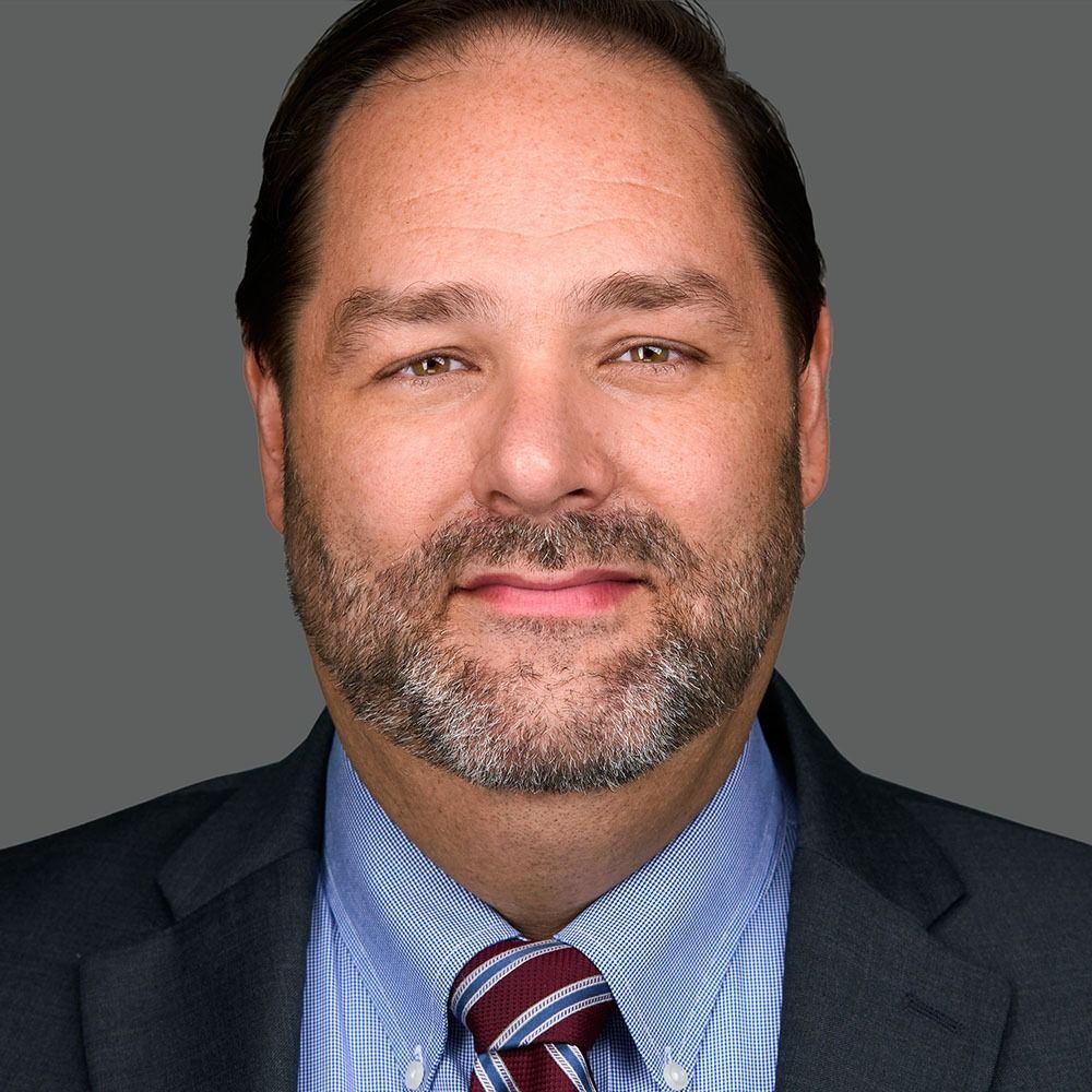 Man in a suit, light blue shirt, and patterned tie; looking at the viewer, neutral expression.