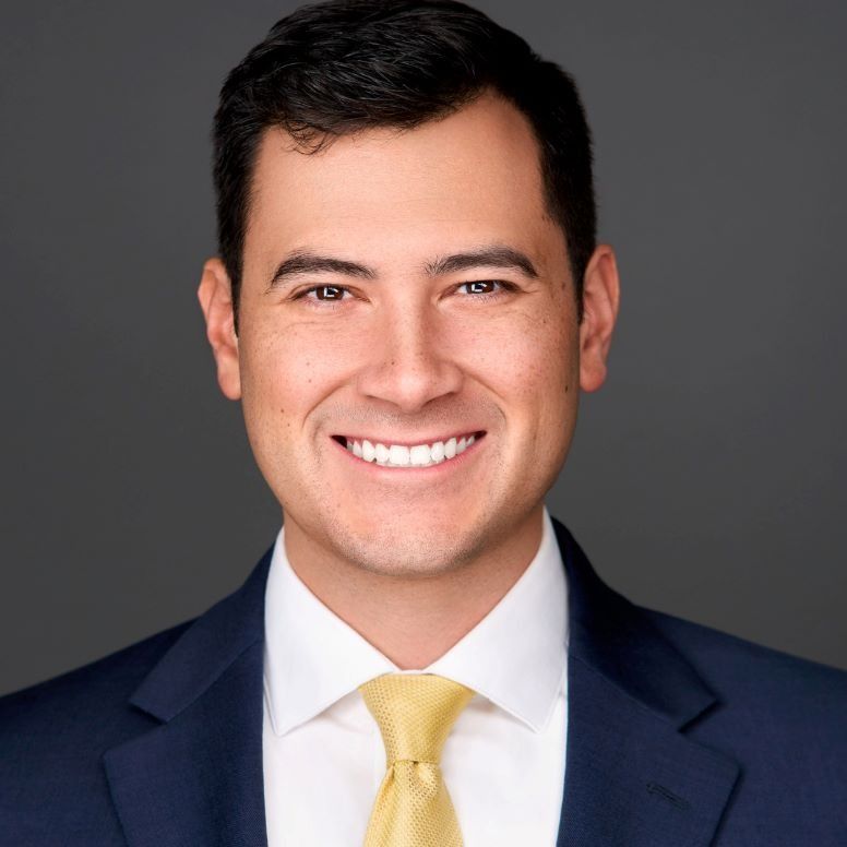 Man in a navy suit and yellow tie smiles at the camera against a gray backdrop.