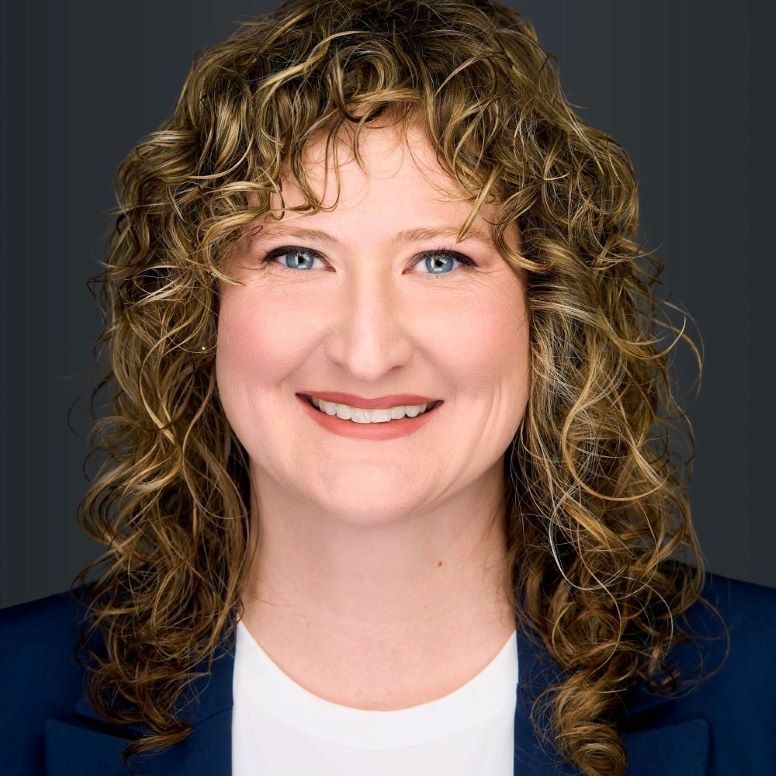 Woman with curly brown hair, smiling, wearing a blue blazer and white top.