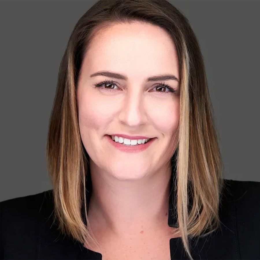 Woman with brown hair smiling, wearing a black blazer, against a gray background.