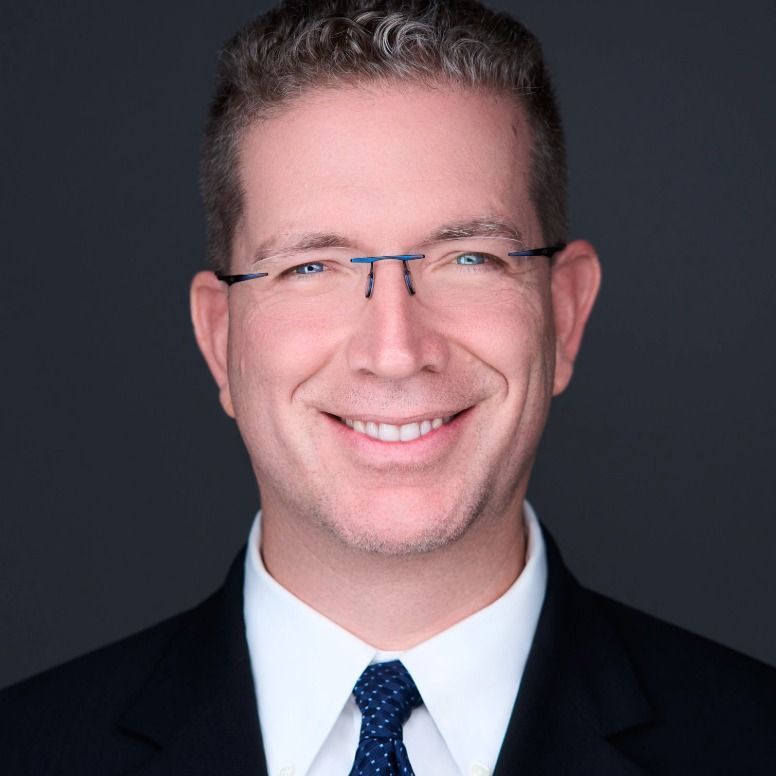 Man with glasses, smiling, wearing a suit and tie, against a gray background.