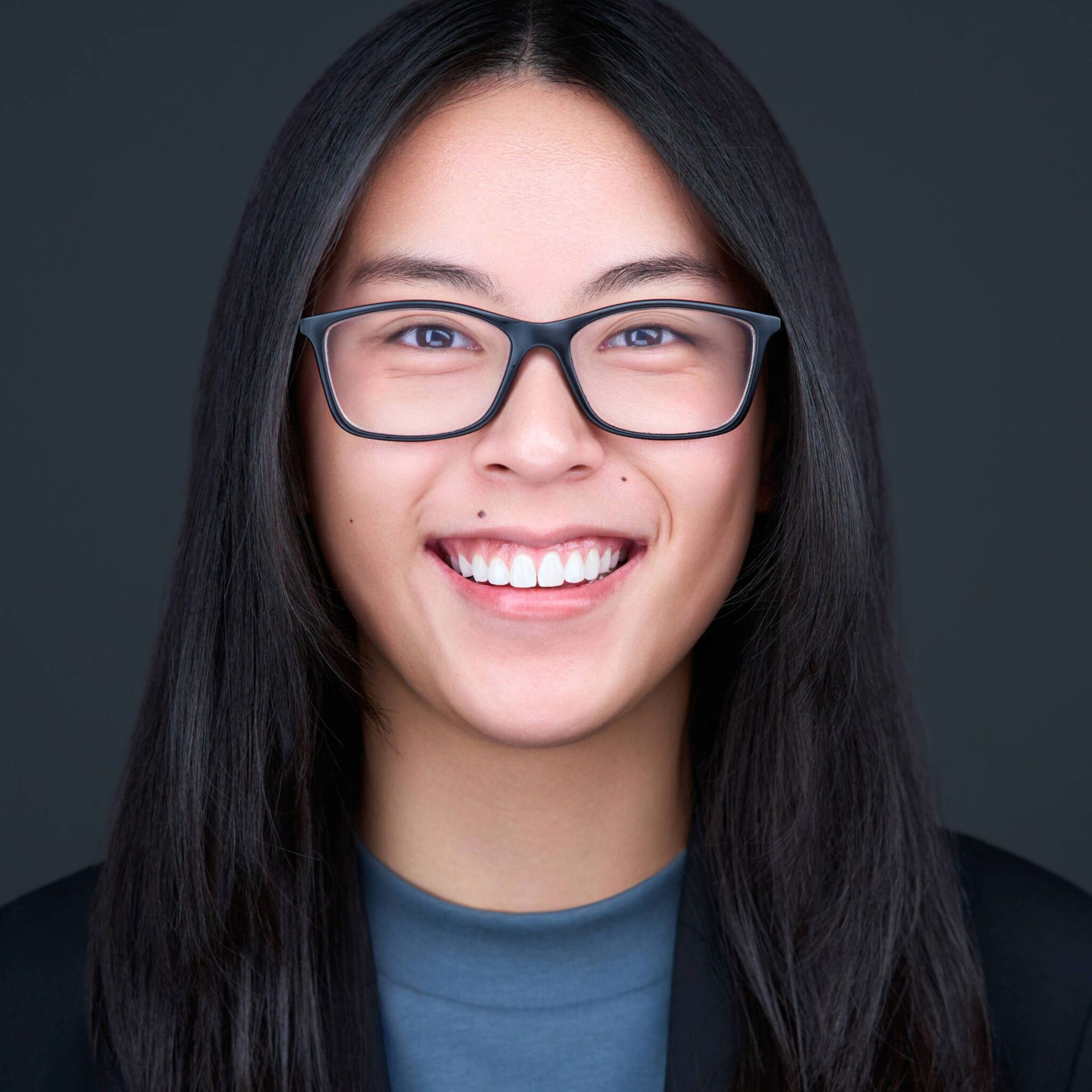 Woman with glasses smiling, looking at the camera against a dark gray background.