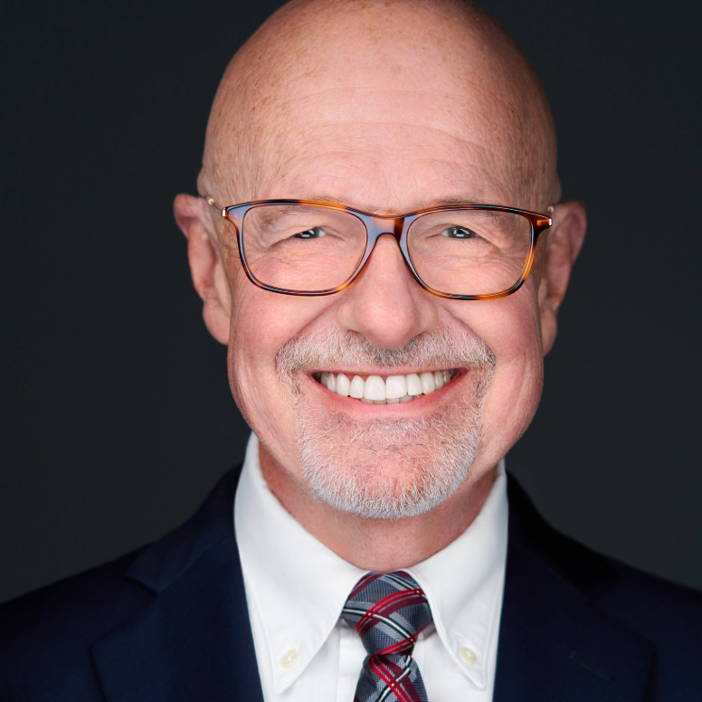 Bald man with glasses smiles, wearing a navy suit and patterned tie, against a dark gray background.