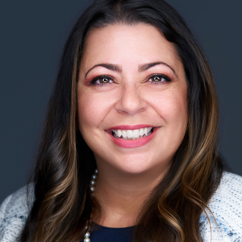 Woman with long brown hair smiles, wearing a pearl necklace and a white jacket, against a dark background.