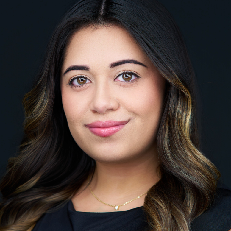 Woman with dark hair smiles, gold necklace, dark background.