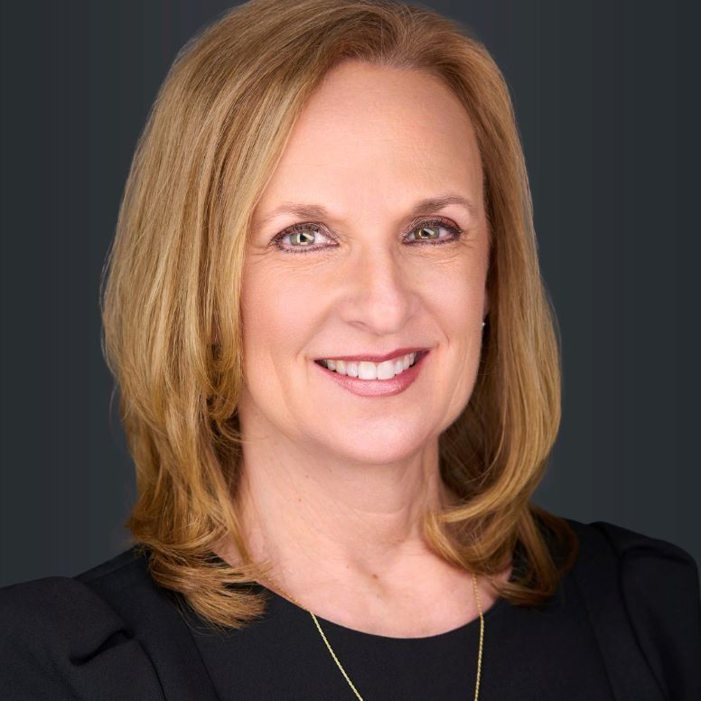 Woman with blonde hair smiling, wearing a black top and gold necklace against a dark background.