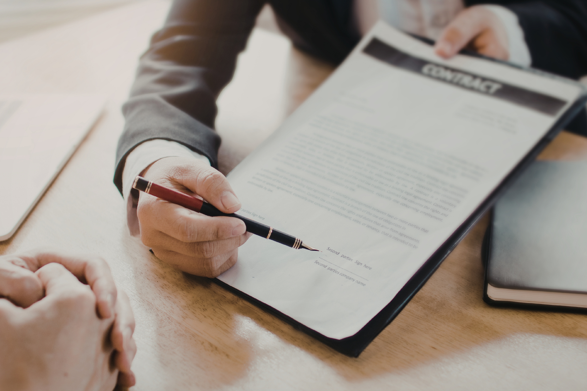 Person in suit signing a contract with a pen, sitting at a wooden table.