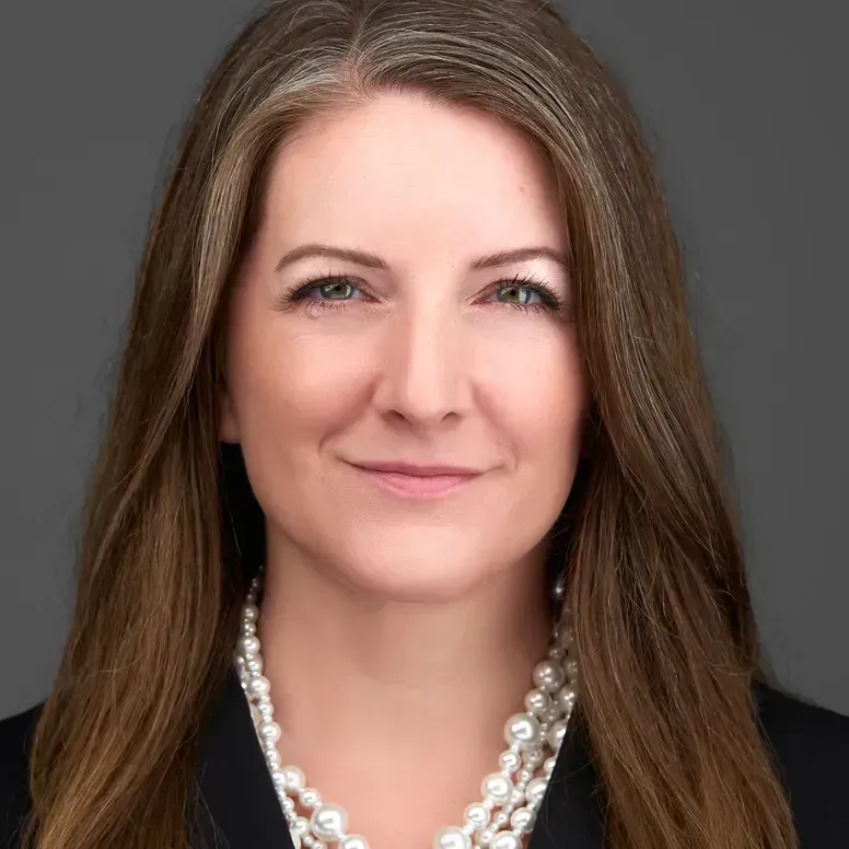 Woman with long brown hair wearing a pearl necklace and black blazer, smiling.