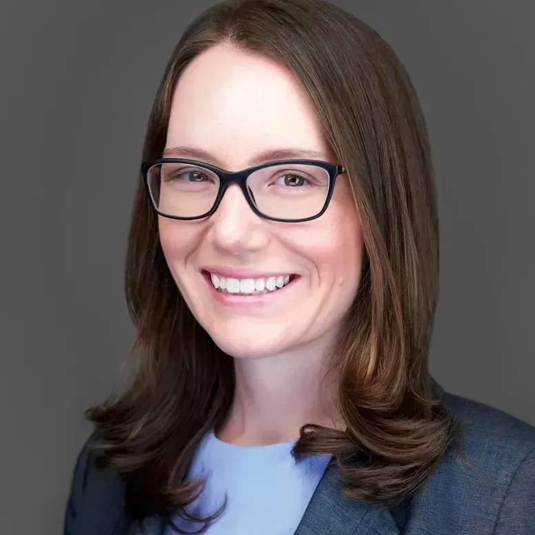Woman with glasses smiles, wearing a blue shirt and grey blazer, against a grey background.