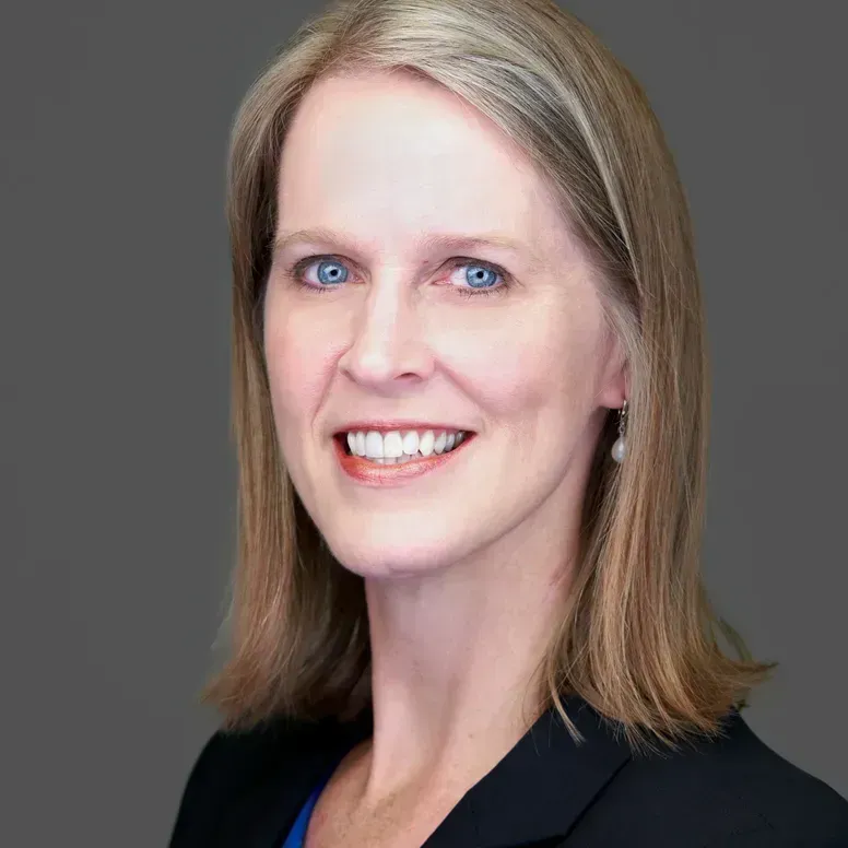 Woman with fair skin and blue eyes smiles at the camera, wearing a dark blazer, pearl earrings, against a gray backdrop.