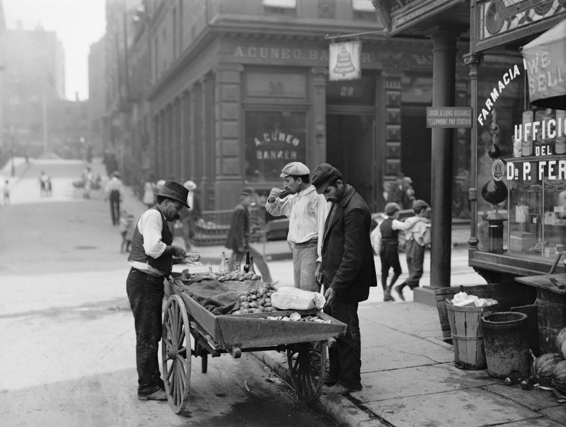 A black and white photo of a man standing next to a cart on a city street.