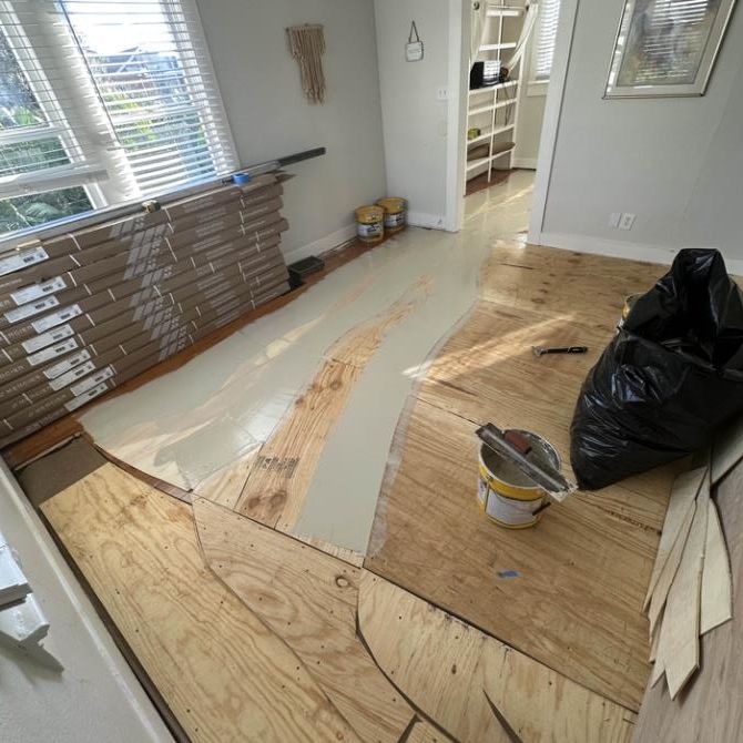 Room with unfinished wood flooring, partially covered with gray adhesive, with new flooring materials stacked nearby.