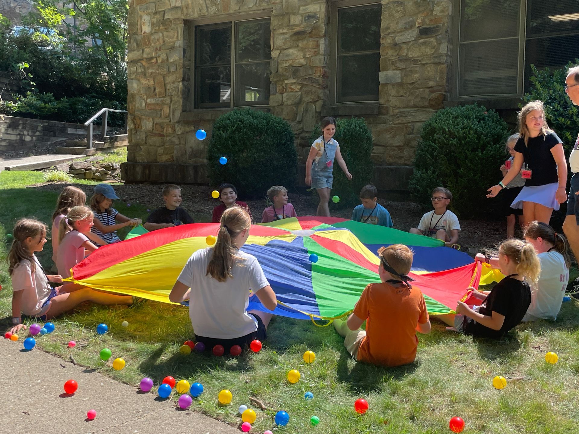 Children and adults play parachute games on a lawn outside a stone building with colorful balls scattered around.