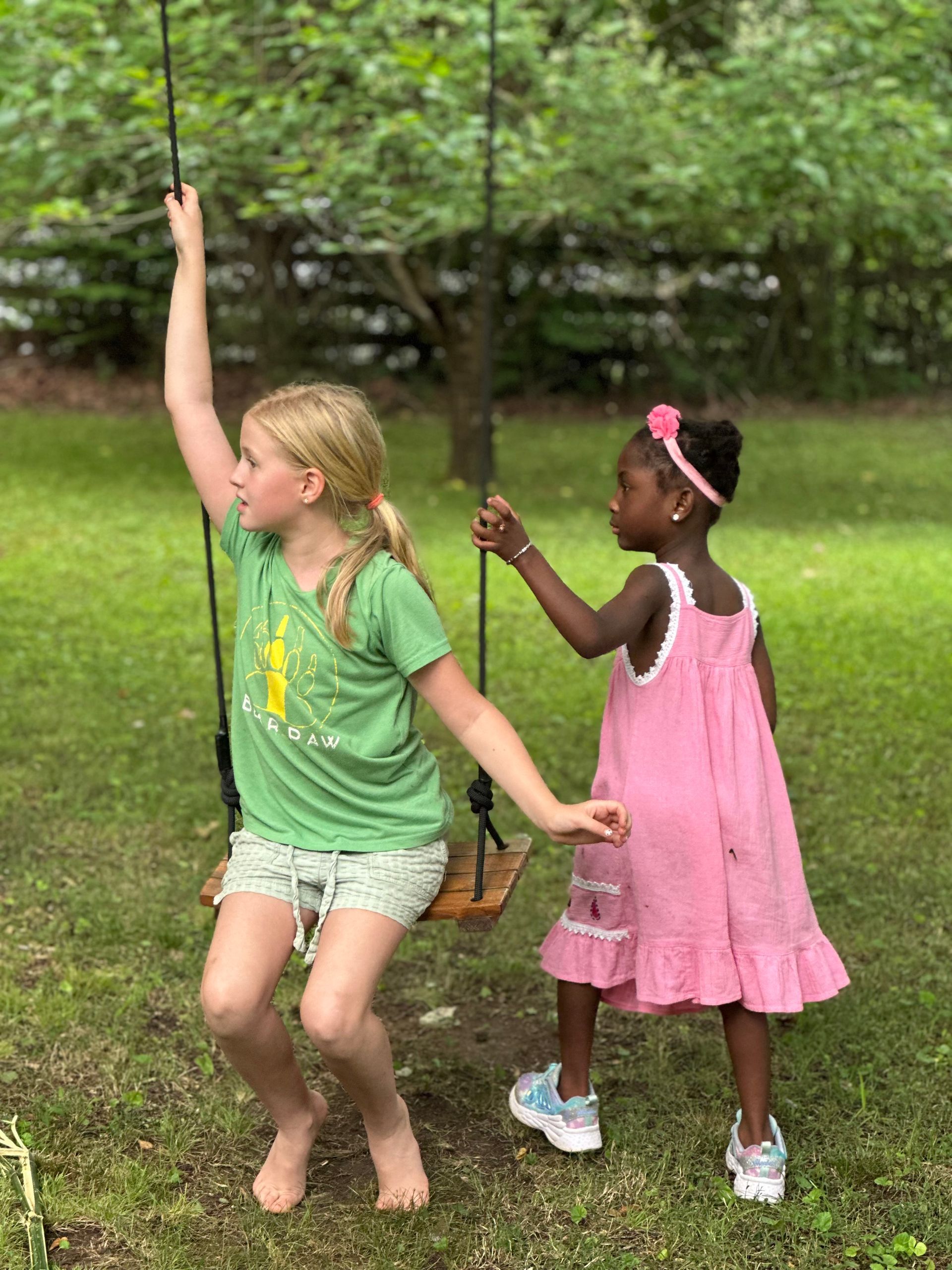 Two girls play outside on a swing, one with a green t-shirt sitting on the swing and one in a pink dress ready to assist.