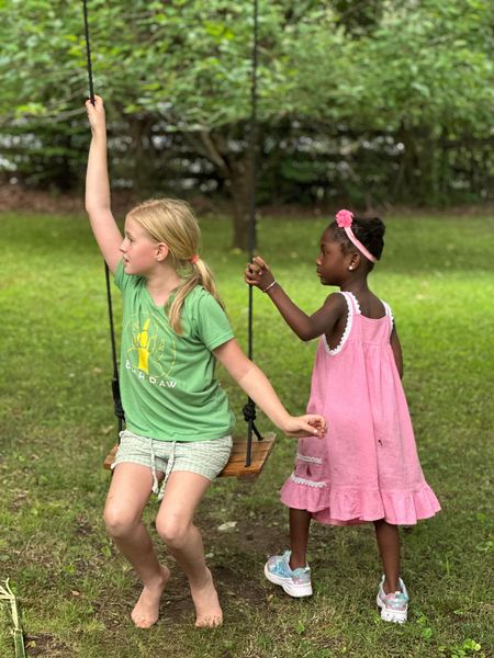 Two girls play outside on a swing, one with a green t-shirt sitting on the swing and one in a pink dress ready to assist.