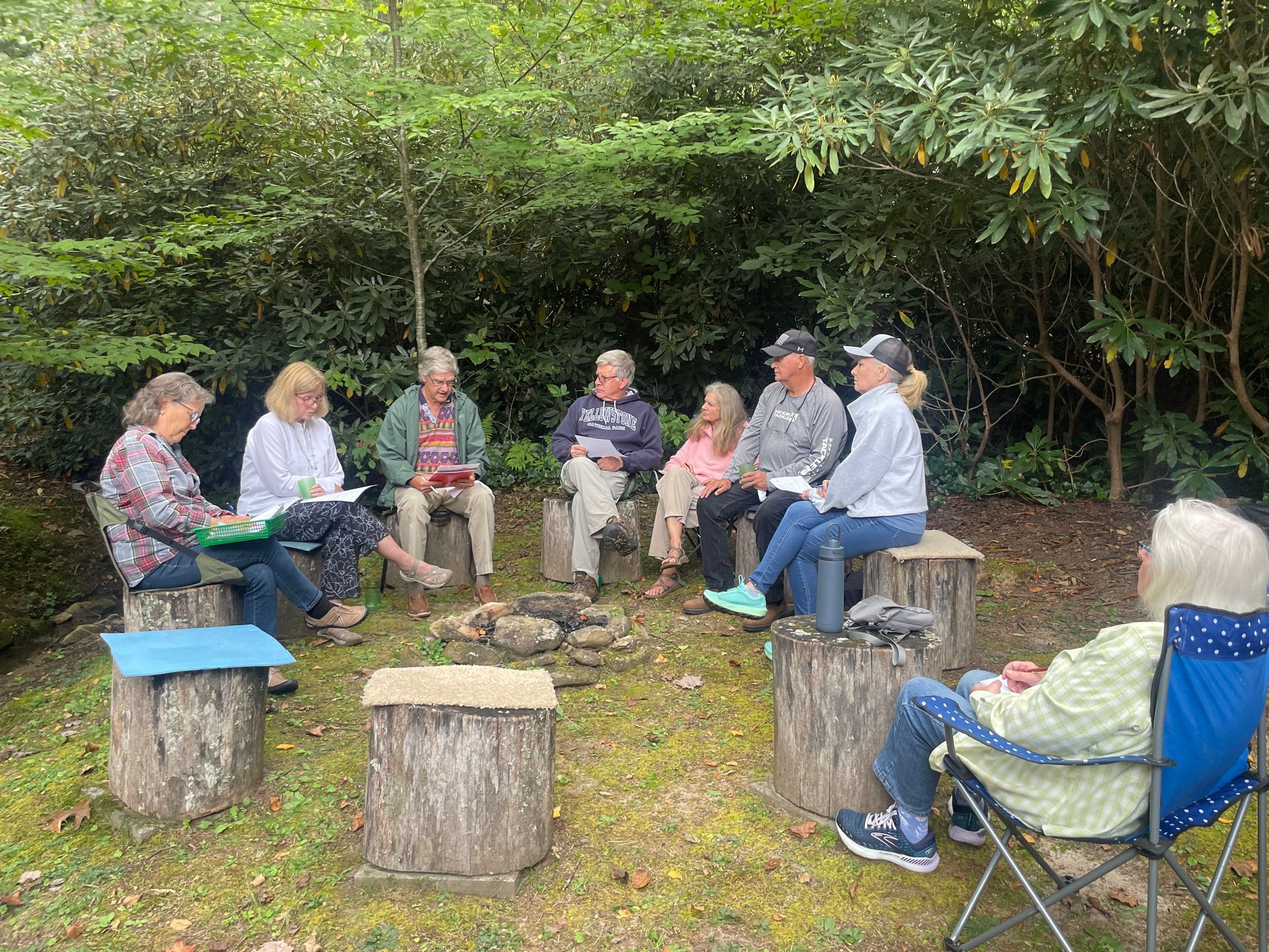 People sitting in a circle on tree stumps outdoors in a wooded clearing, having a group discussion.
