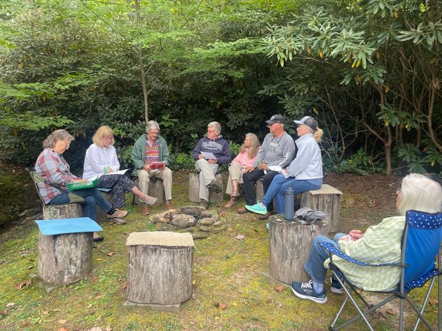 People sitting in a circle on tree stumps outdoors in a wooded clearing, having a group discussion.