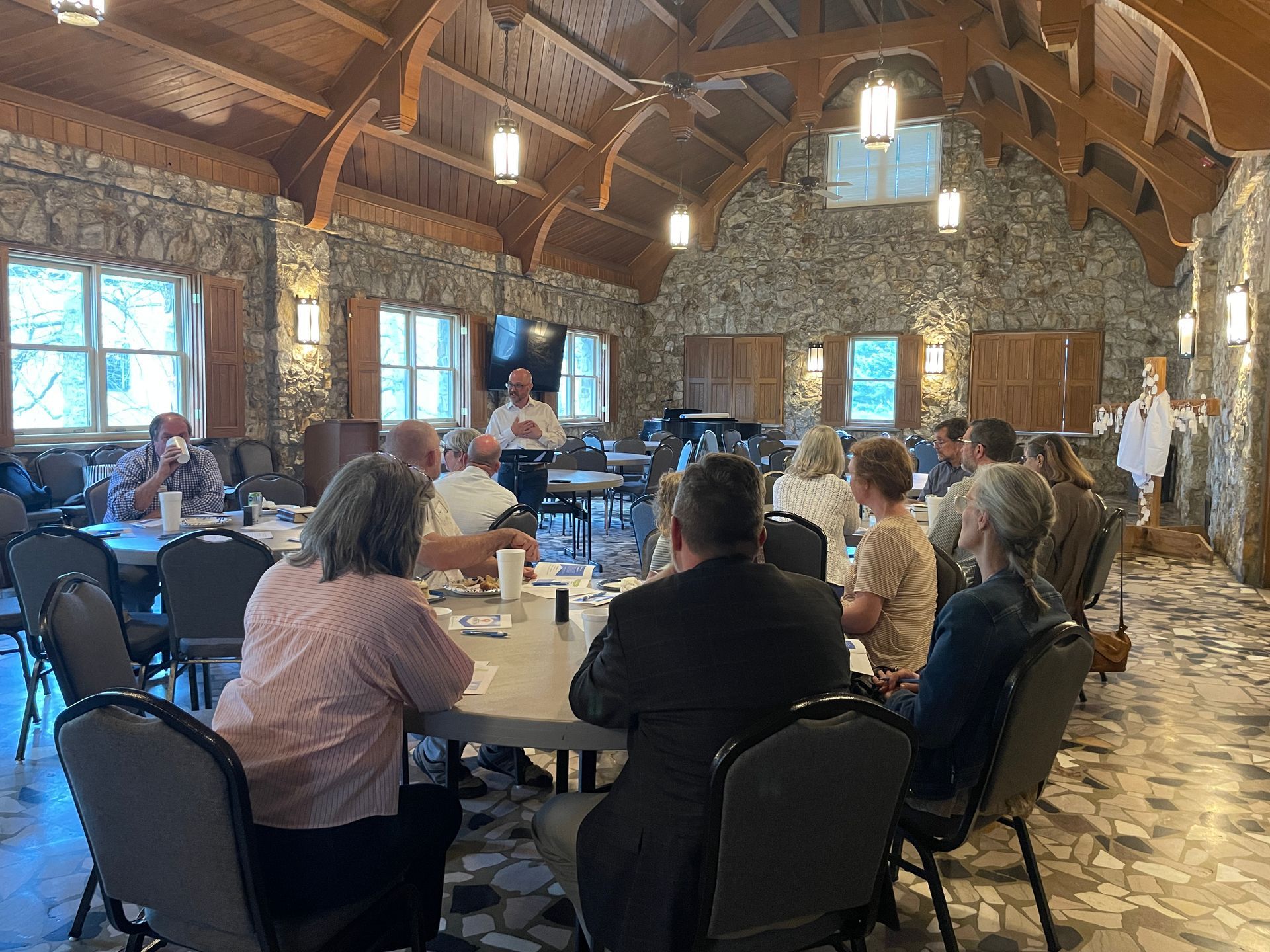 People seated around round tables in a rustic stone hall with wood beams and hanging lights.
