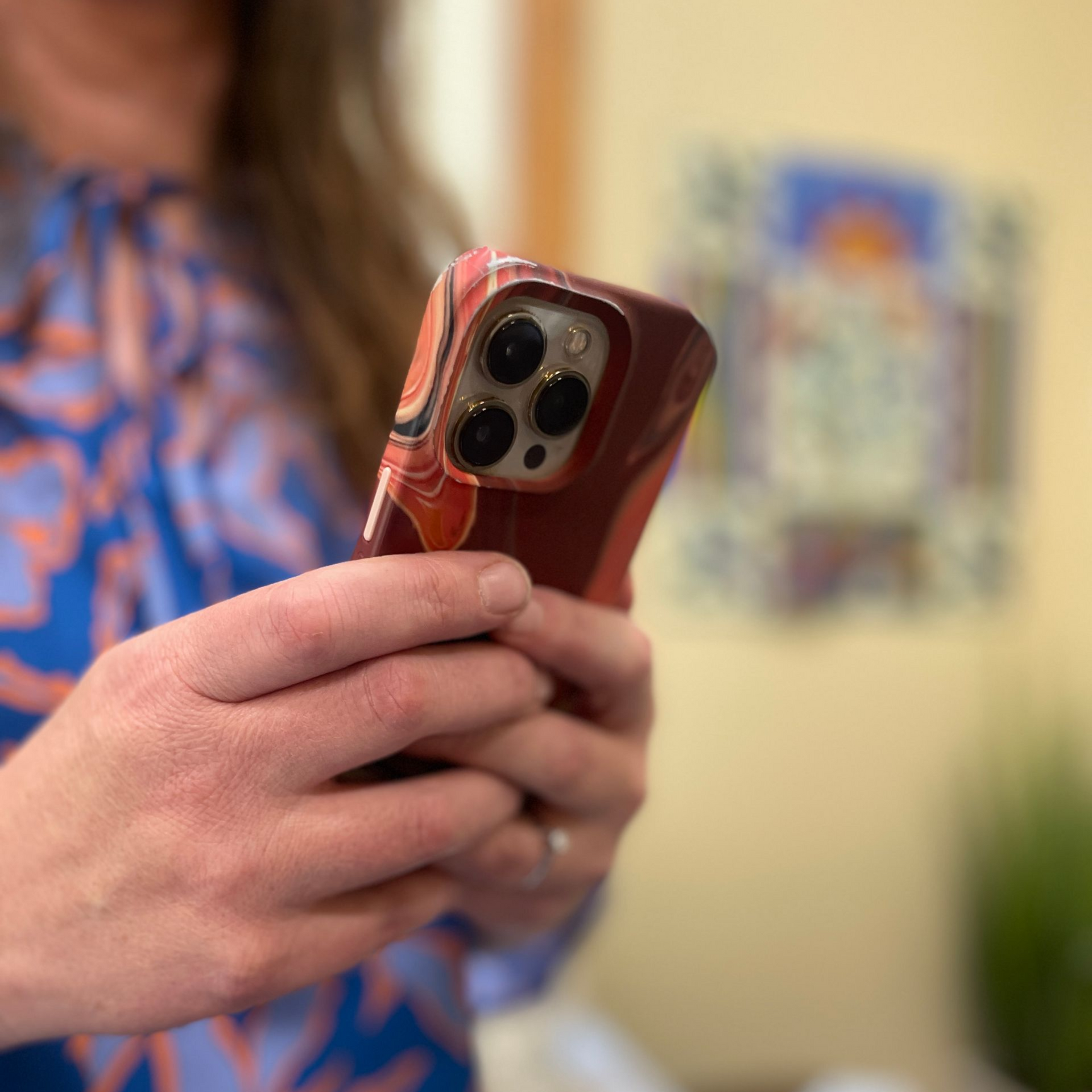Hands holding a red smartphone with a triple camera case in a blurred indoor setting