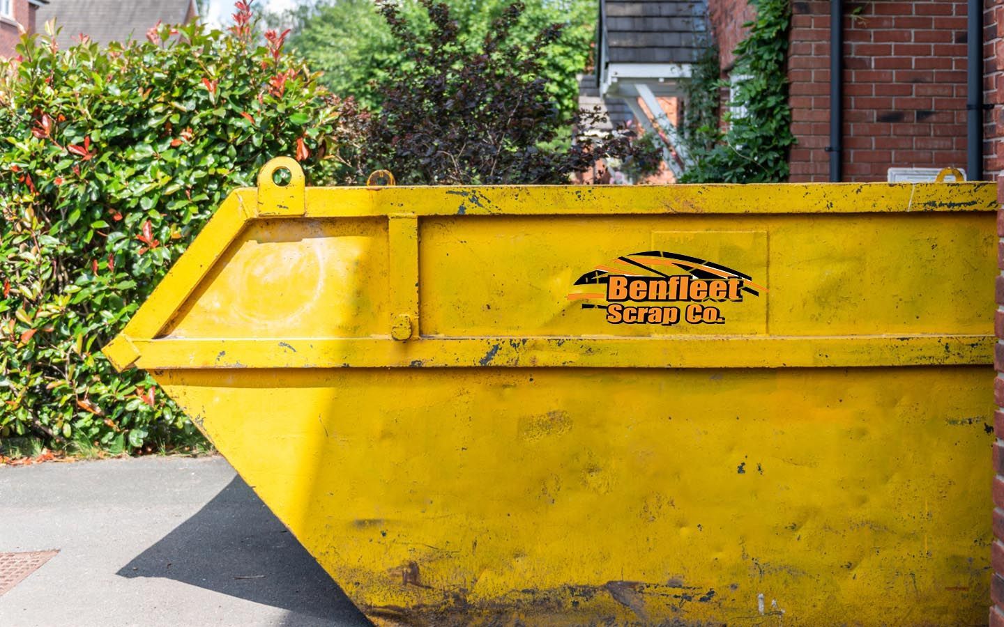 A yellow skip is parked in front of a brick building with Benfleet Scrap logo