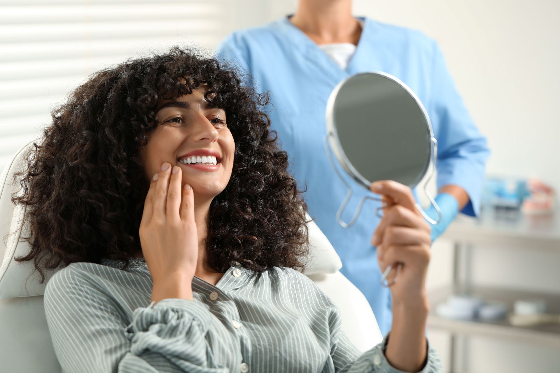 Woman smiles, examining her teeth in a hand mirror at a dental office, with a dental professional in the background.