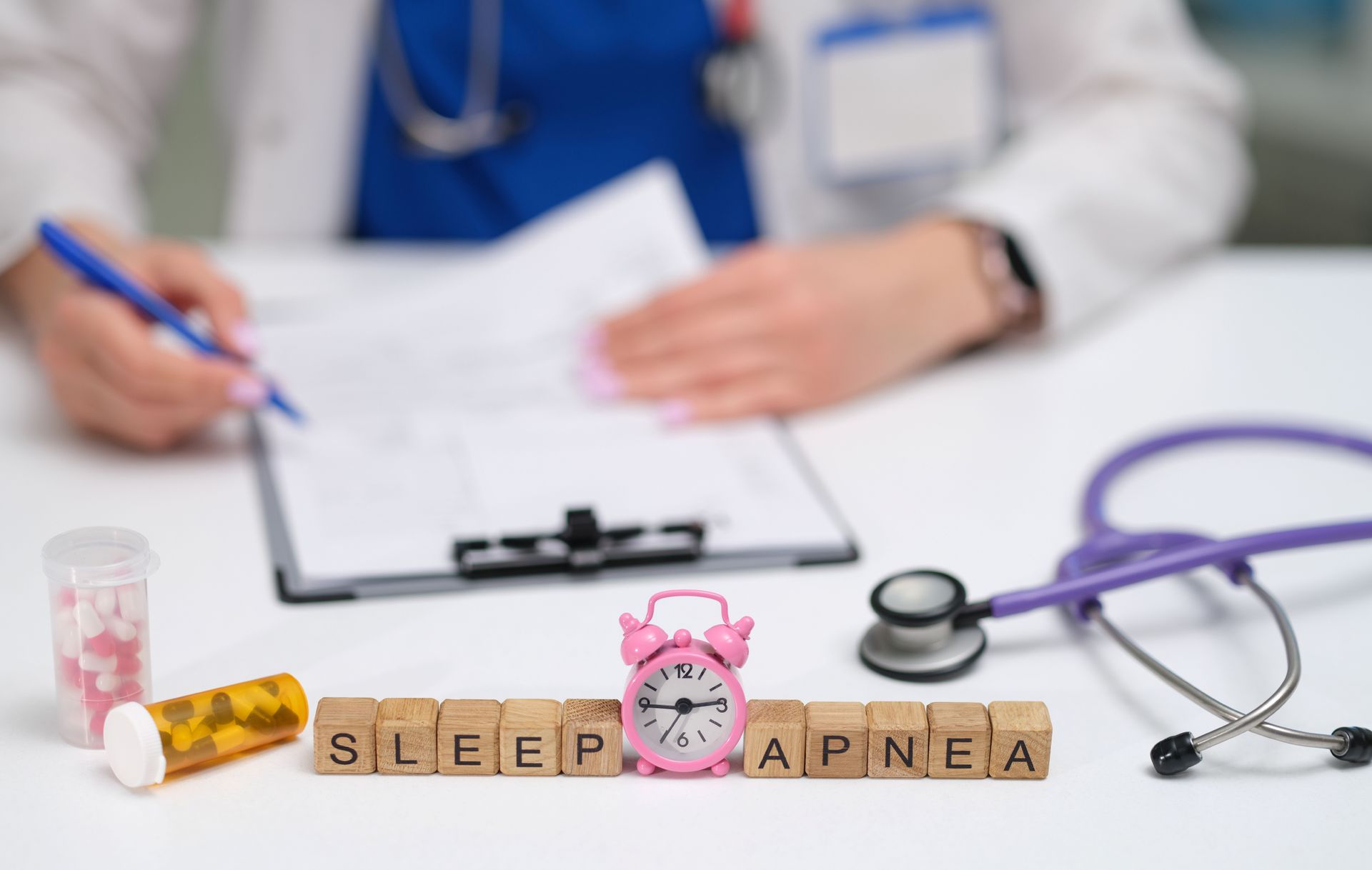 A medical professional writing on a clipboard; sleep apnea spelled out with letter blocks, pills, alarm clock, stethoscope.