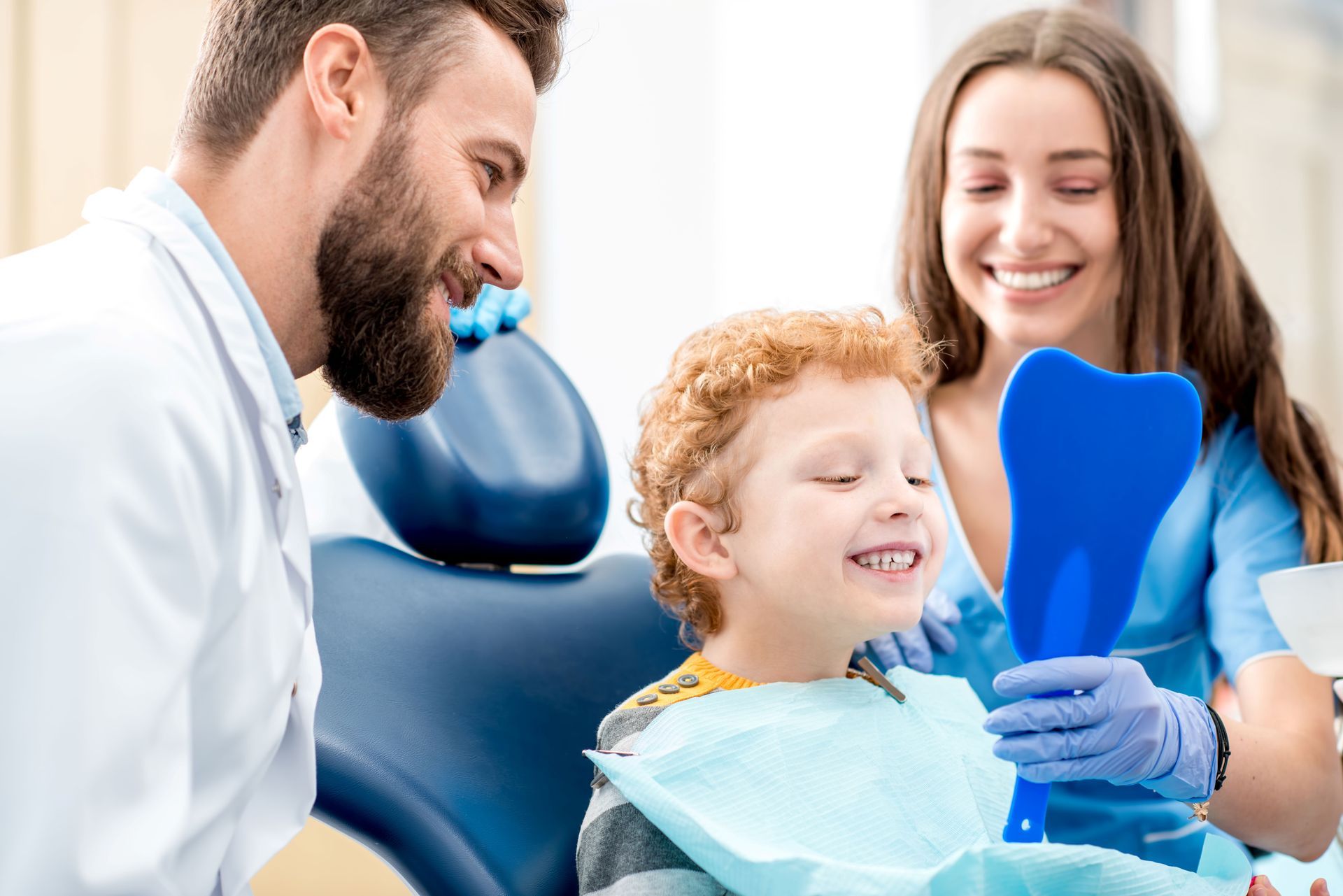 Boy looking in a mirror at teeth, with dentist and dental assistant smiling in a dental office.