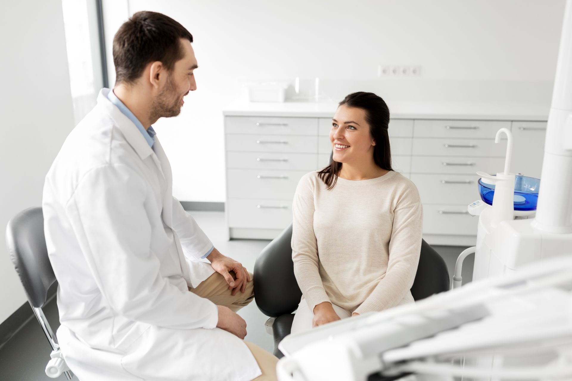 Dentist in white coat talking to a smiling patient seated in a dental chair in a bright office.