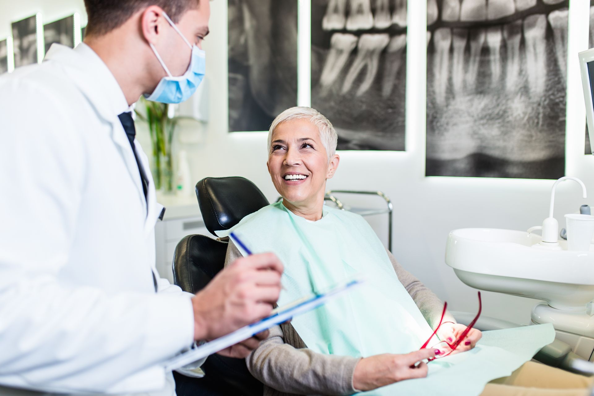 Dentist in mask reviews paperwork with patient in dental chair, holding glasses; x-rays on wall.