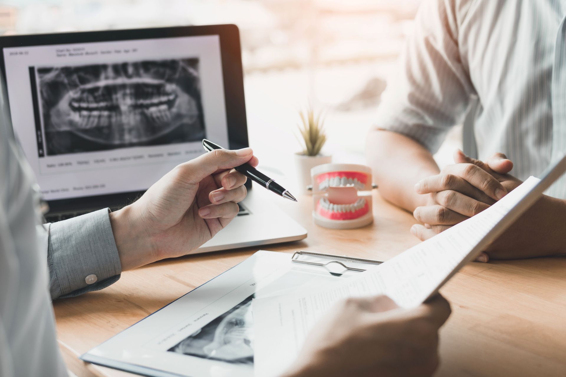 Dentist pointing at x-ray on laptop, explaining to patient. Model teeth and paperwork on table.