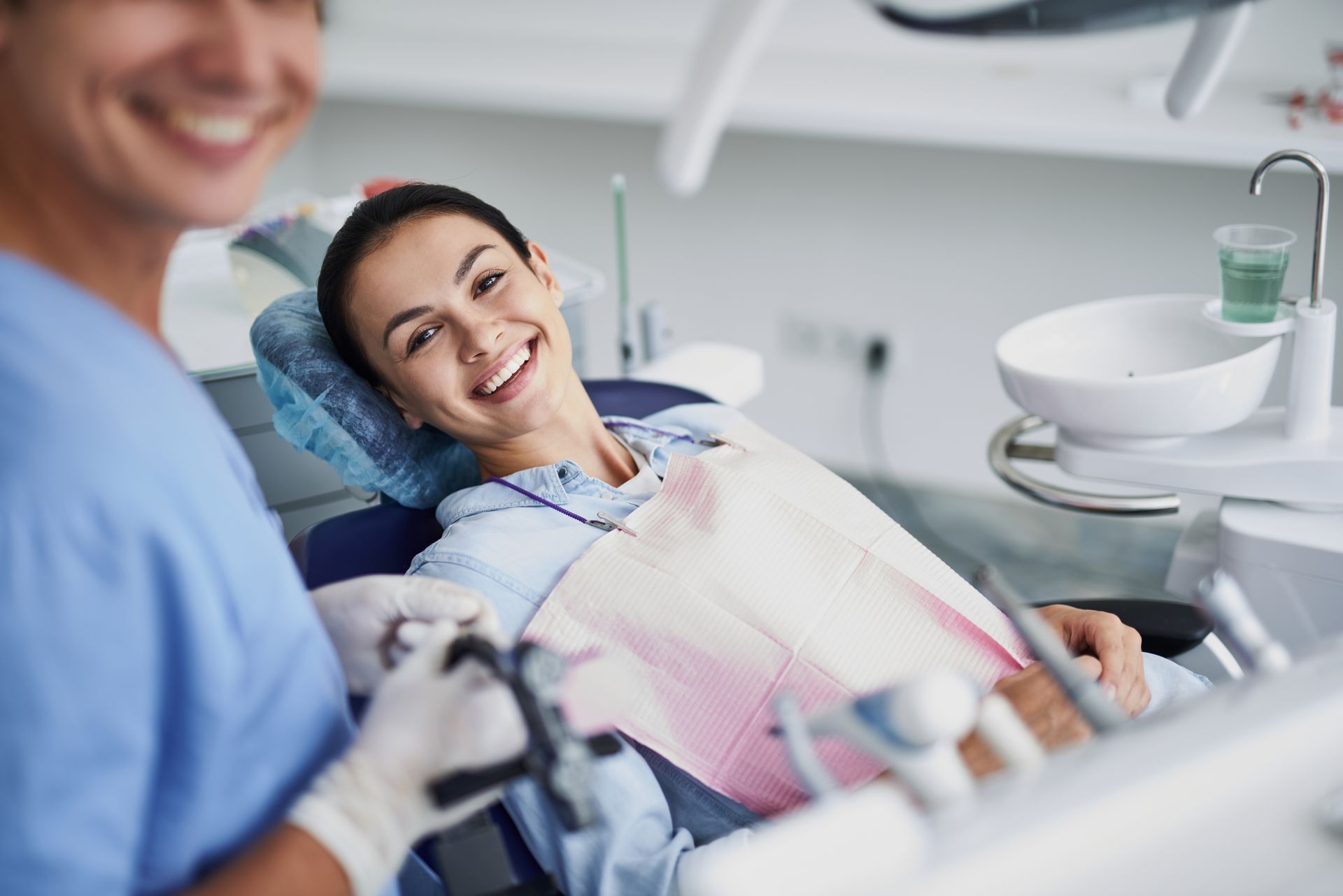 Woman smiles in a dentist's chair; a dentist is partially visible, also smiling. Dental equipment in the background.