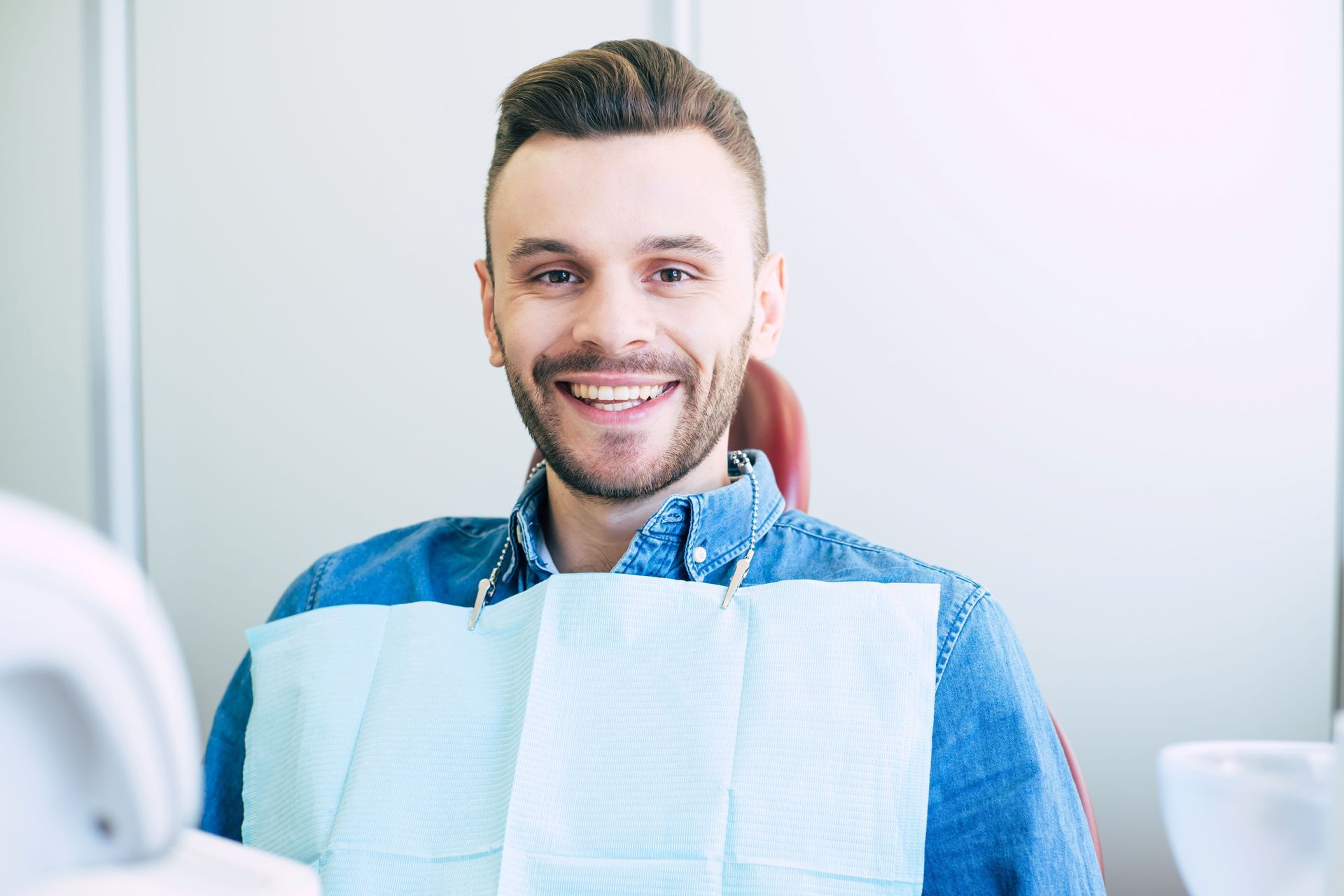 Man smiles in a dentist's chair, wearing a bib, inside a dental office.