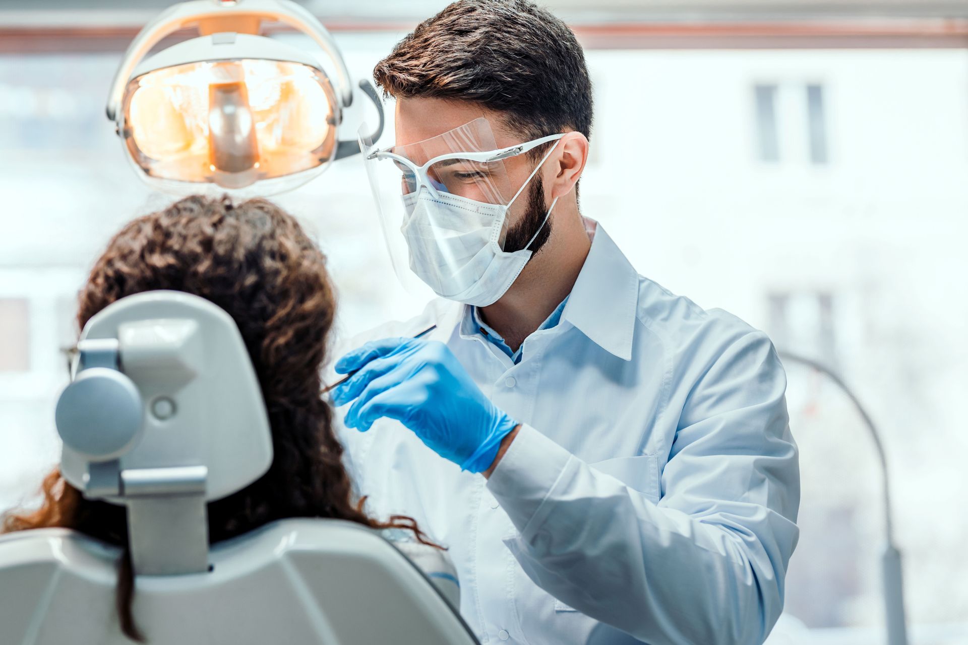 Dentist examining a patient's mouth. The dentist wears a mask and shield, uses a tool, and blue gloves.
