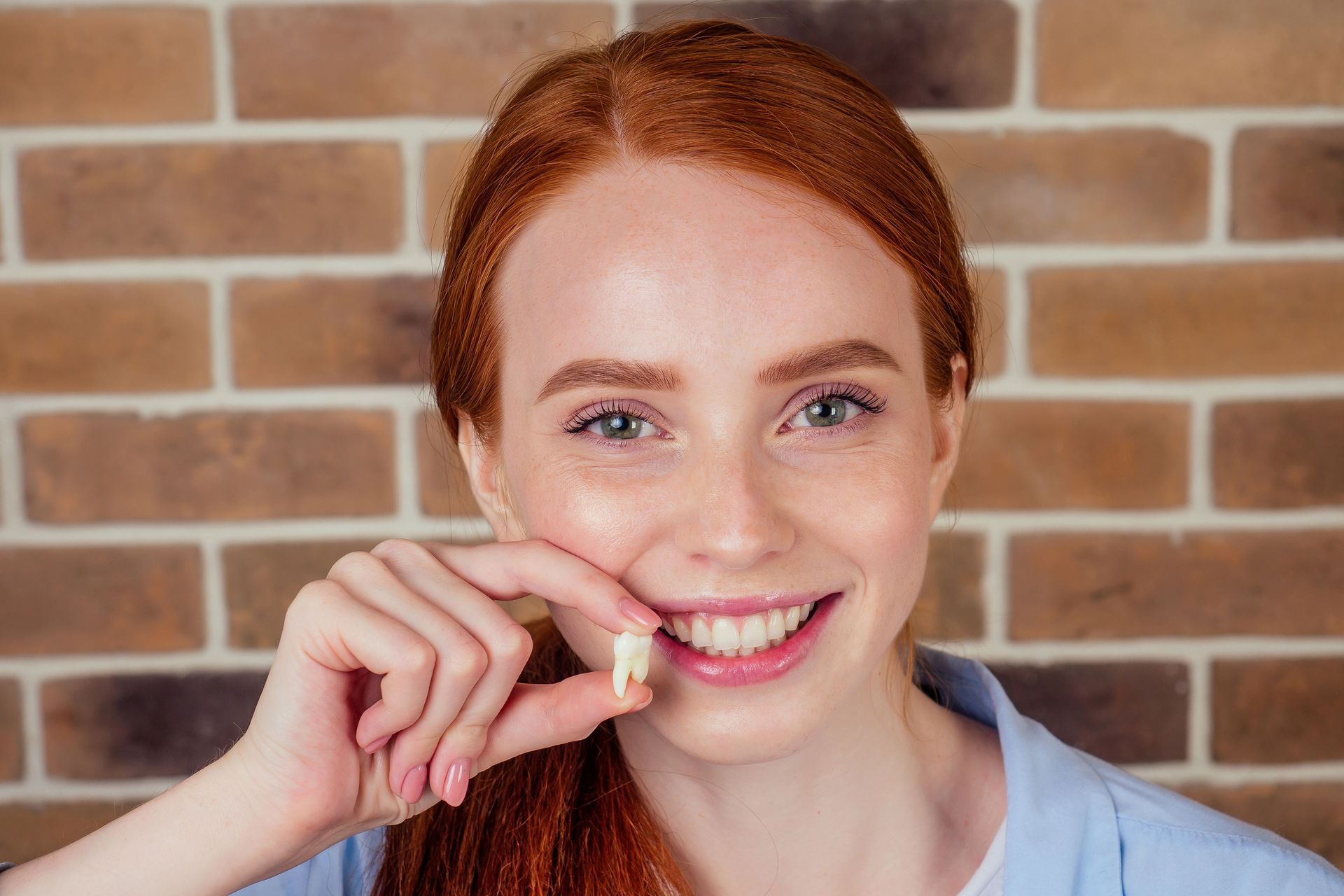 Woman with red hair smiling, holding a tooth in front of a brick wall.