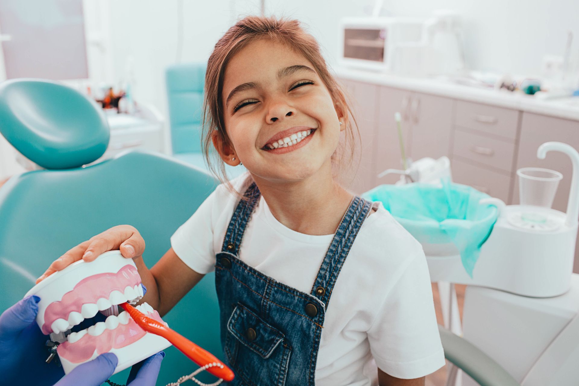 Girl smiles, holding model teeth with a toothbrush, in a dentist's office.