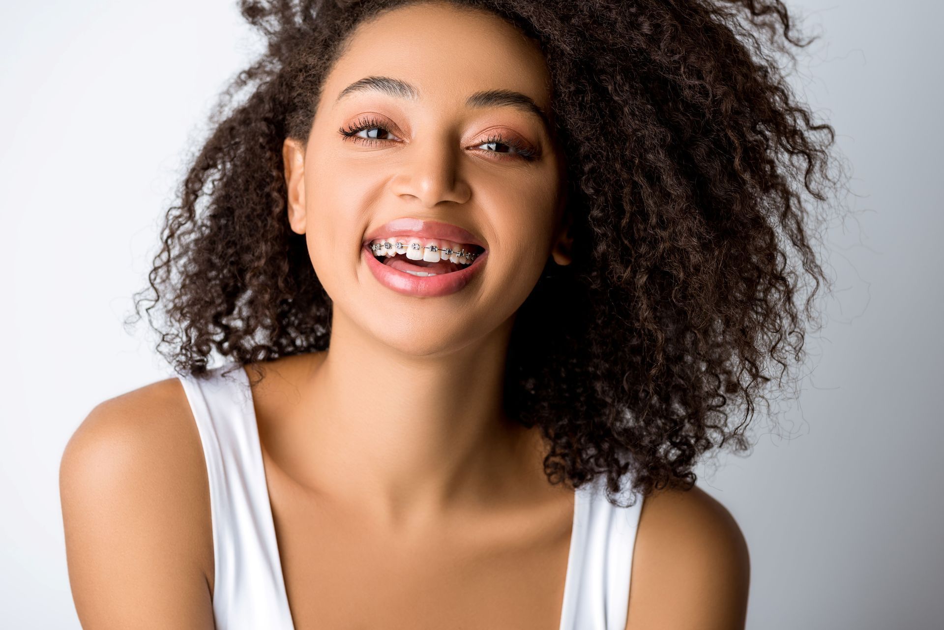 Person with curly hair smiles, showing braces, white tank top, neutral background.