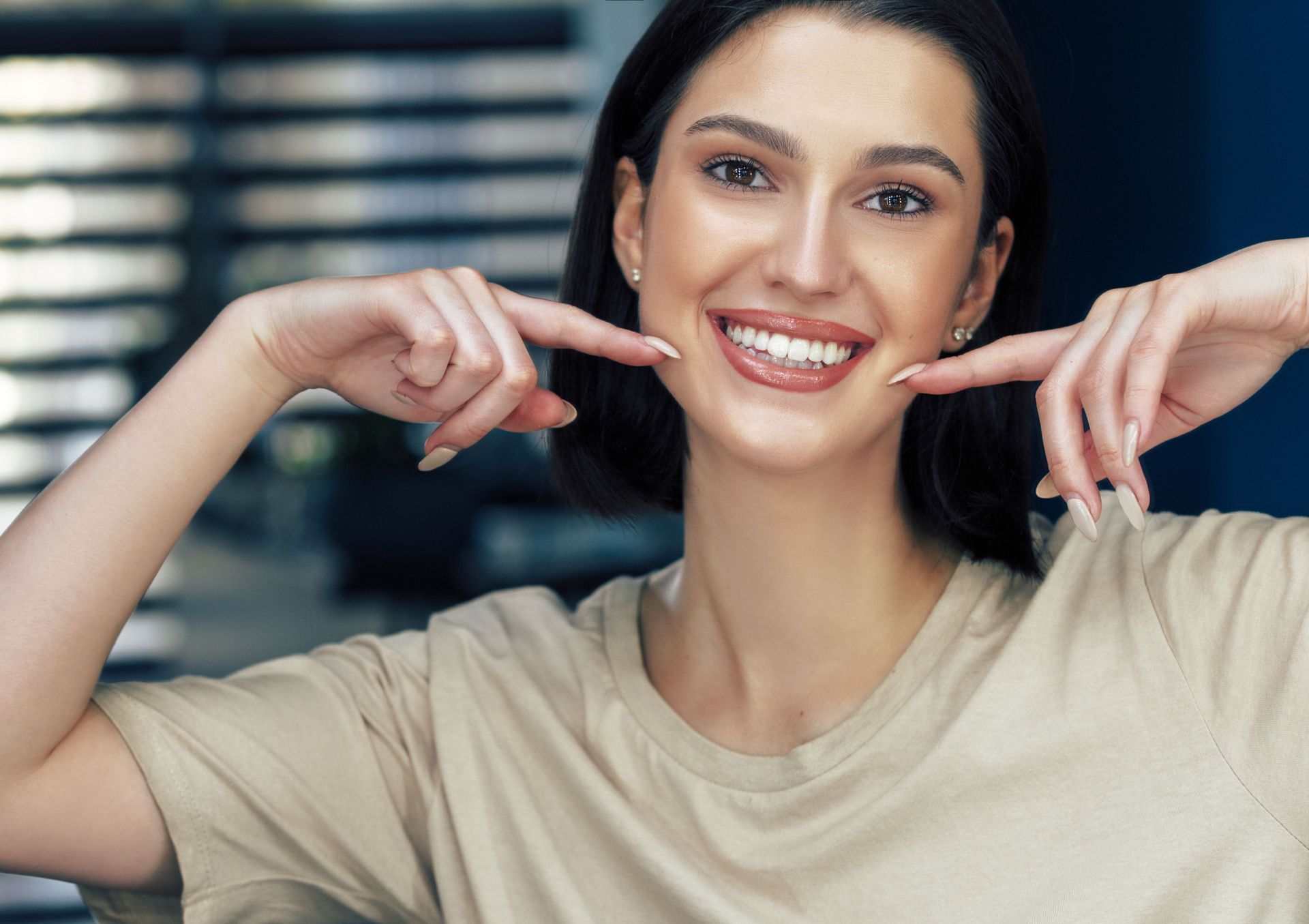 Woman smiling, pointing to her teeth, beige shirt, dark hair, natural light.