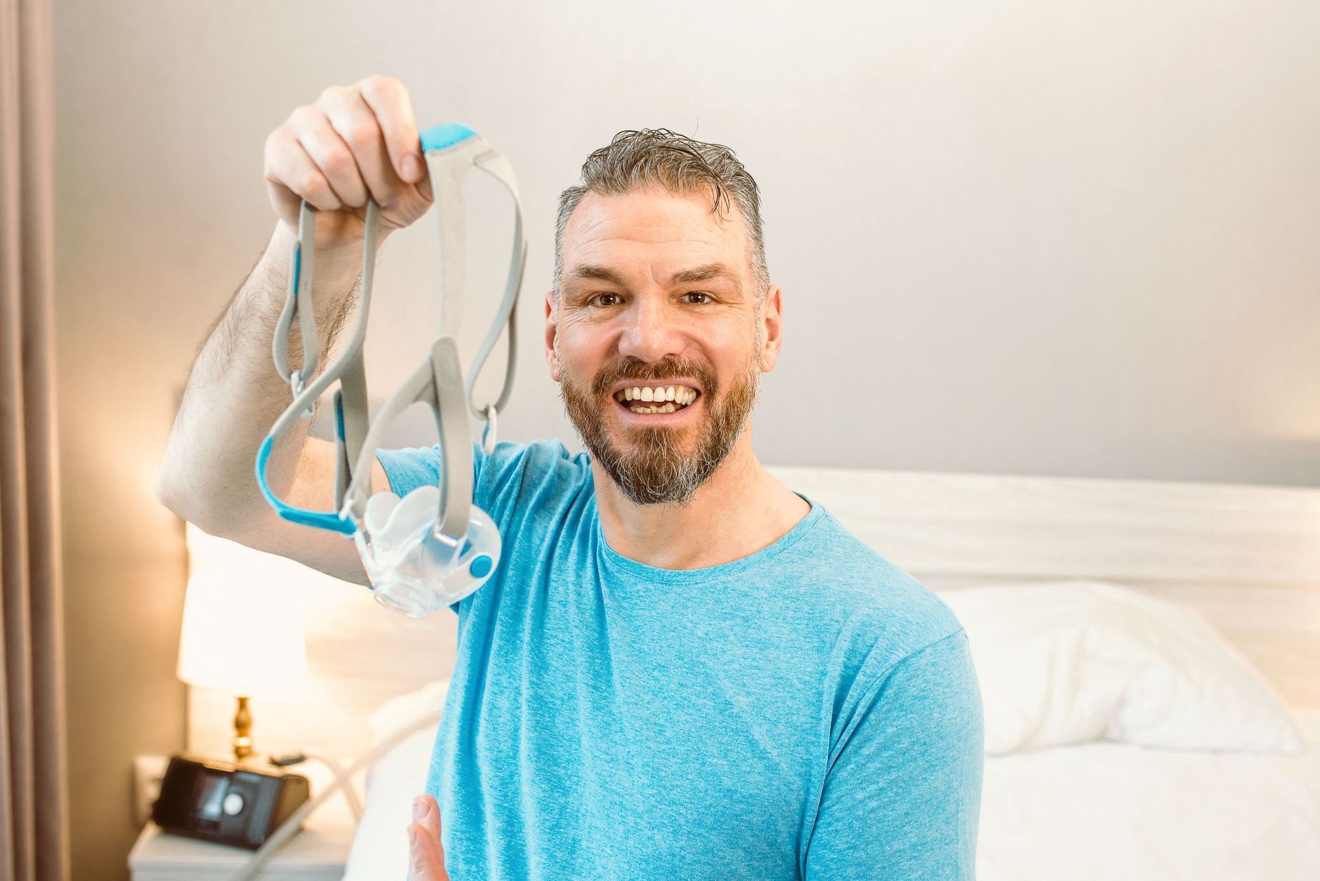 Man holding CPAP mask, smiling in bedroom. Blue shirt, bed in background.