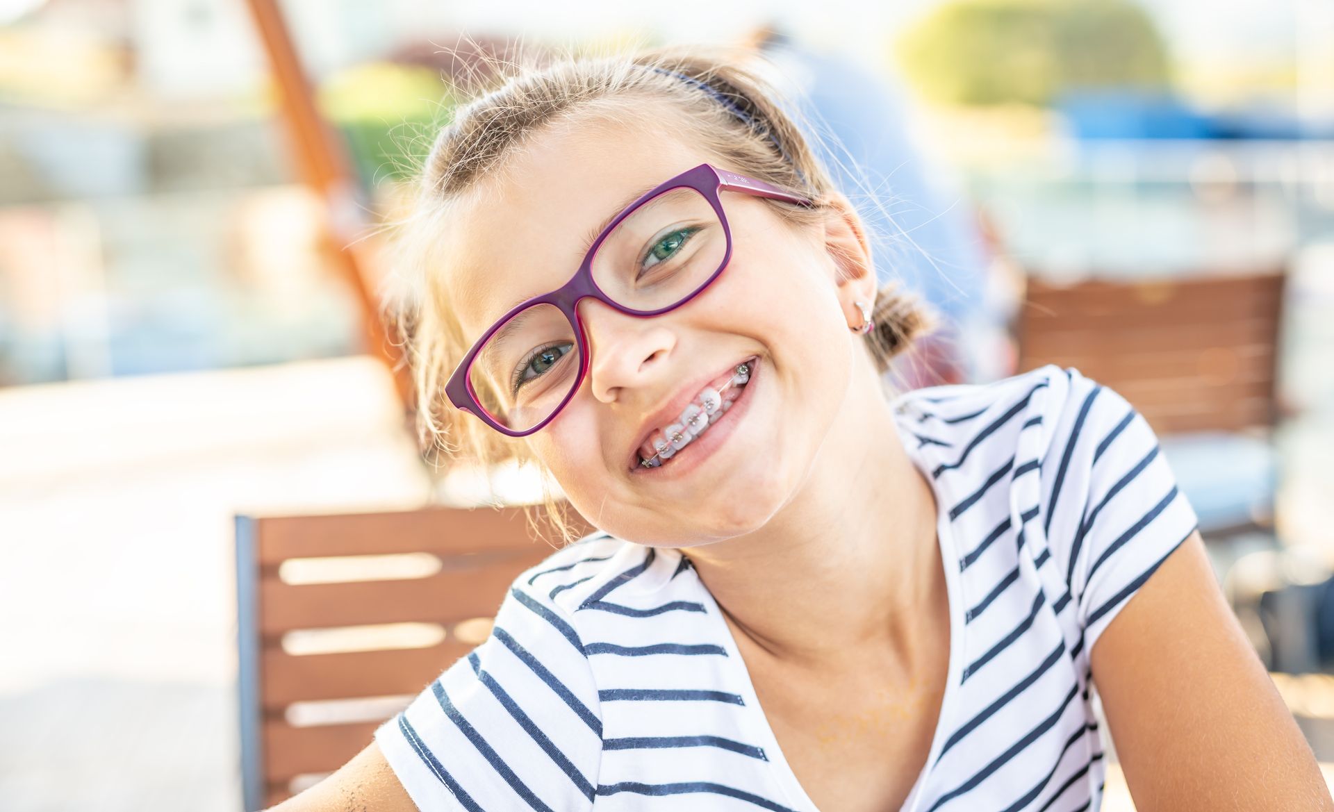 Girl with braces and glasses smiles, wearing striped shirt, seated outdoors.
