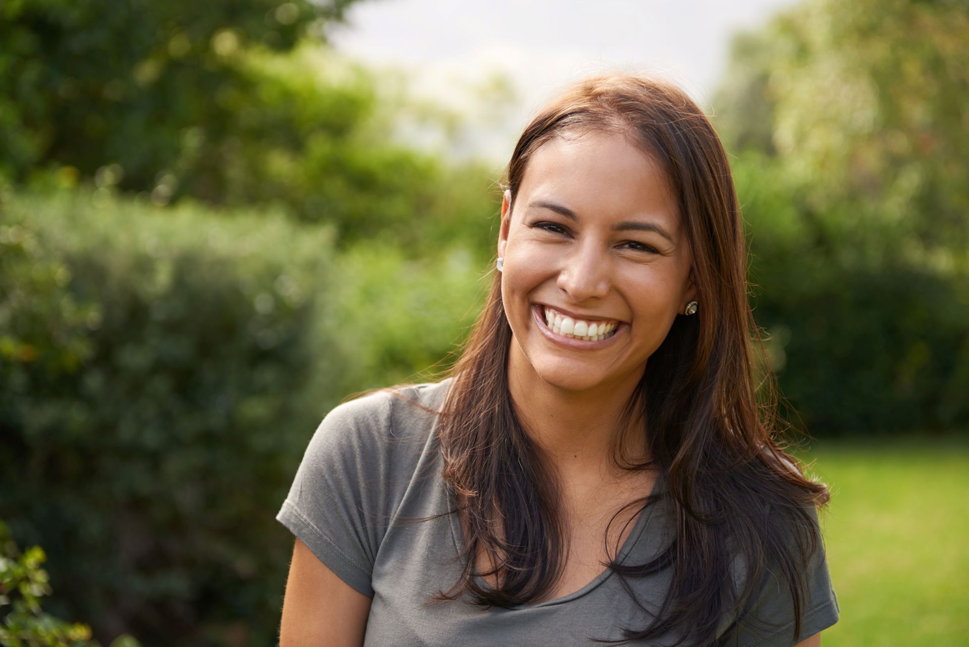 Woman smiling outdoors in front of green foliage; wearing a gray t-shirt.