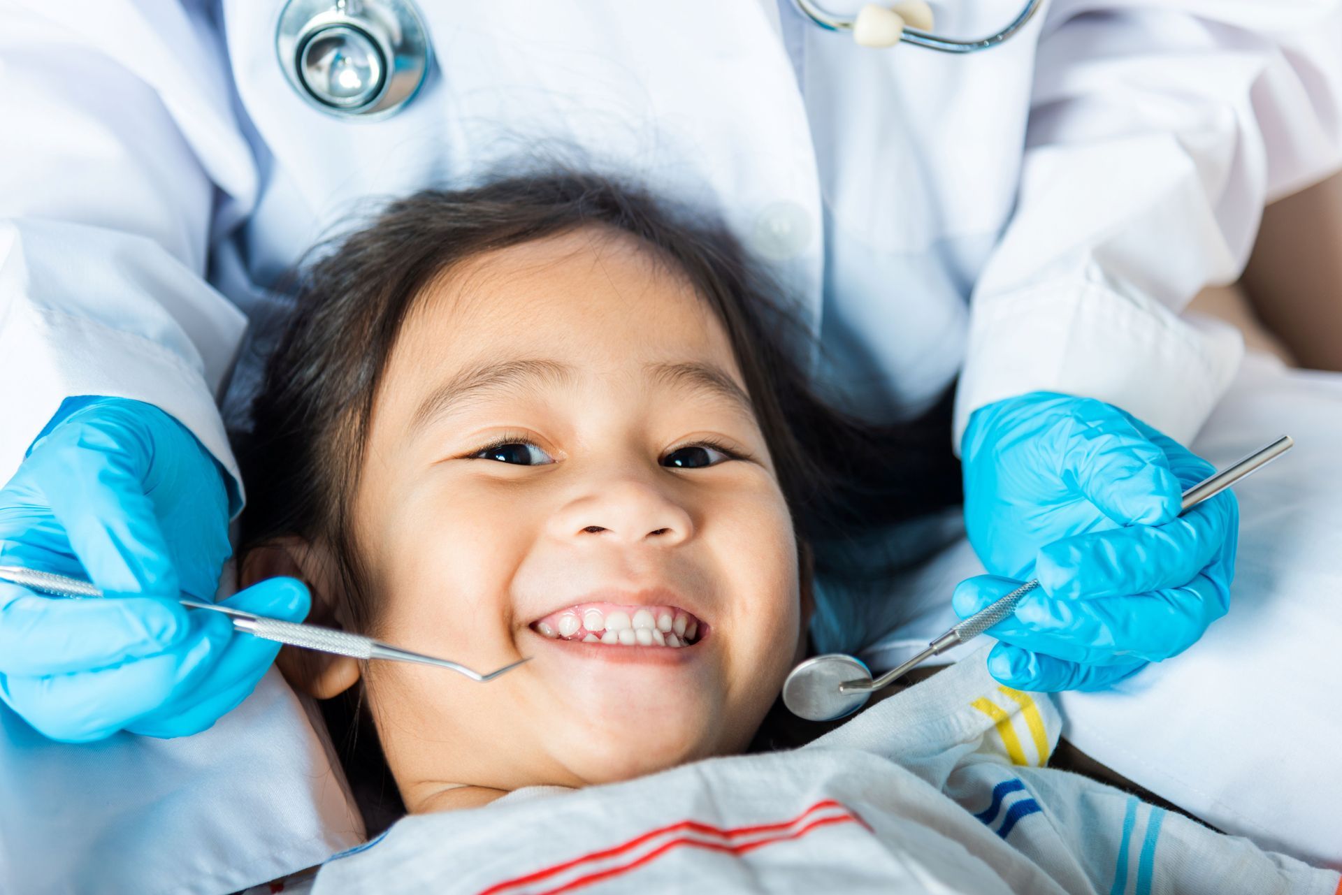 Young child smiles during dental checkup, dentist in gloves holds tools.