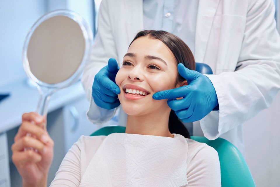 Dentist examining a patient's teeth. Patient is smiling, holding a mirror. Blue gloves. Dental chair.