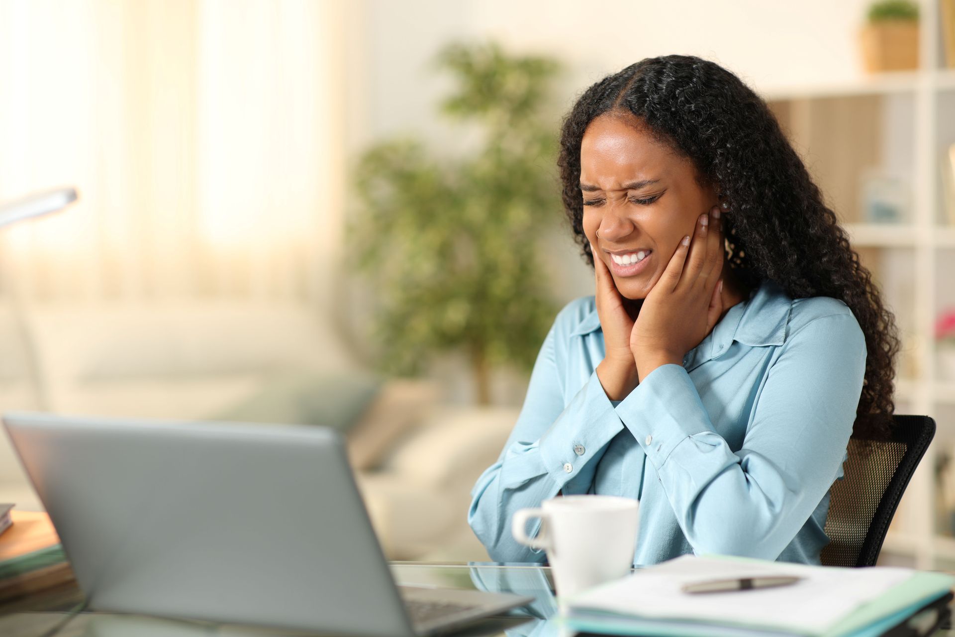 Woman at desk with laptop, grimacing while holding cheeks, possibly in pain.