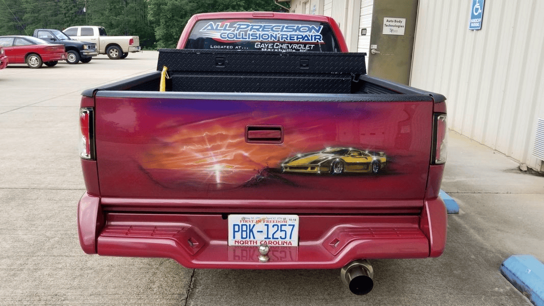 A red truck with a yellow car painted on the back is parked in a parking lot.