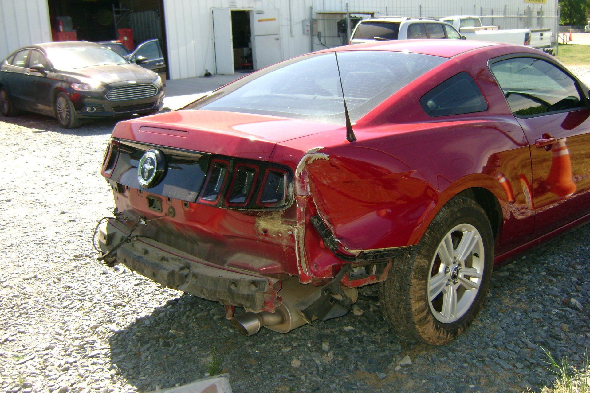 A red ford mustang with a damaged rear end is parked in a gravel lot.